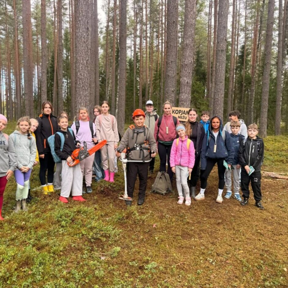Grupa, kurā ir apmēram 15 bērni un daži pieaugušie, stāv priežu mežā uz sūnu klātas zemes. Viens zēns oranžā ķiverē tur krūmgriezi, bet cits bērns – rotaļu motorzāģi. Fonā koka zīme ar uzrakstu 