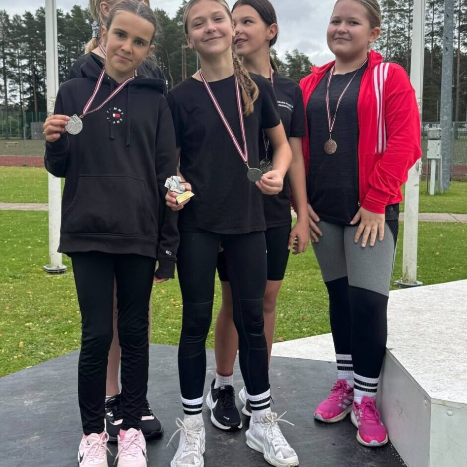 Četras smaidīgas meitenes stāv uz goda pjedestāla ārā, pēc sporta sacensībām. Divas meitenes kreisajā pusē tur sudraba medaļas, un viena vidū tur arī mazu zelta trofeju. Meitenei tālāk pa labi ap kaklu ir bronzas medaļa.