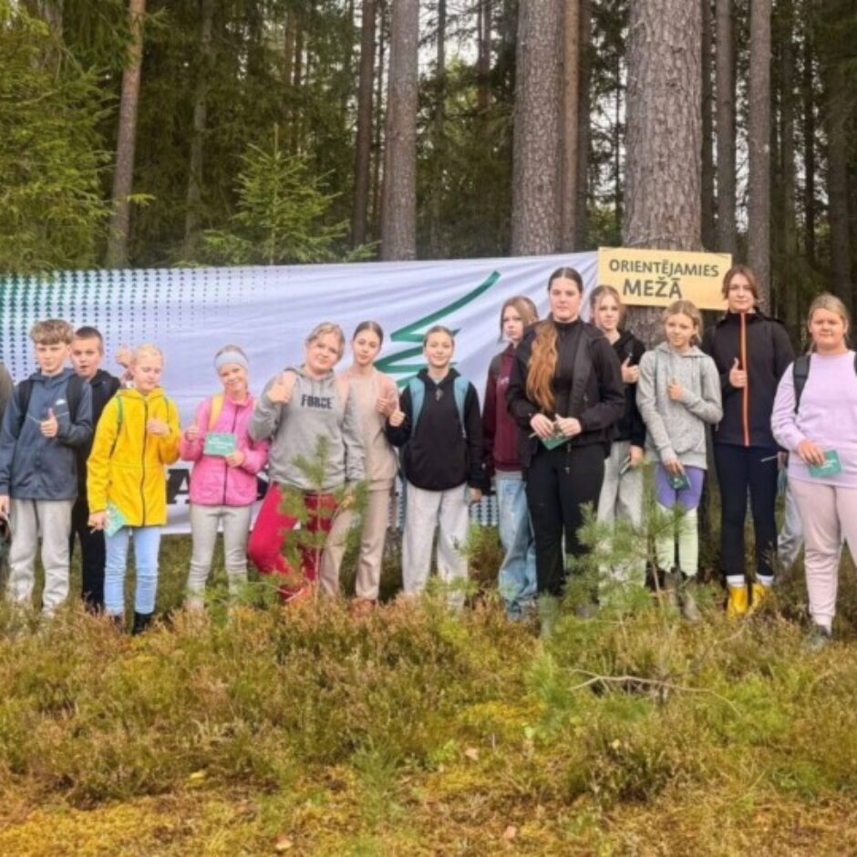 Jauniešu grupa un pieaugusi sieviete stāv mežā uz sūnainas zemes, daudzi rādot īkšķi uz augšu. Fonā redzams balts plakāts ar zaļiem rakstiem un dzeltena zīme uz koka ar uzrakstu 