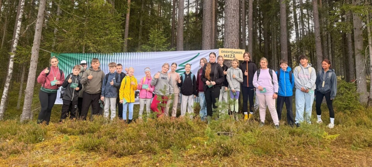 Jauniešu grupa un pieaugusi sieviete stāv mežā uz sūnainas zemes, daudzi rādot īkšķi uz augšu. Fonā redzams balts plakāts ar zaļiem rakstiem un dzeltena zīme uz koka ar uzrakstu 