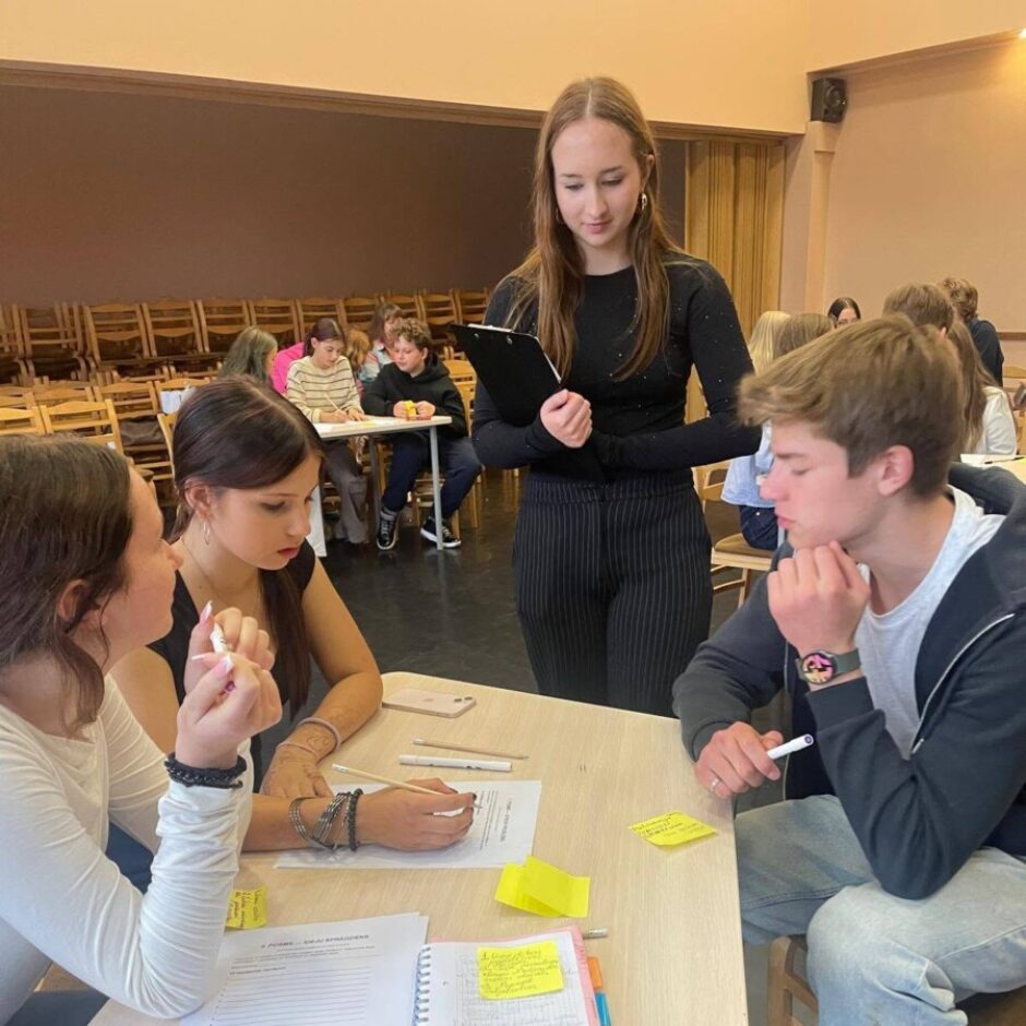 Jauna sieviete melnā apģērbā ar planšeti rokās pārrauga trīs studentus pie galda. Studenti iesaistījušies diskusijā vai uzdevumā; viena meitene raksta, zēns domīgi skatās uz leju, bet vēl viena meitene sarunājas ar kaimiņieni. Uz galda ir dokumenti, tostarp lapa ar virsrakstu 