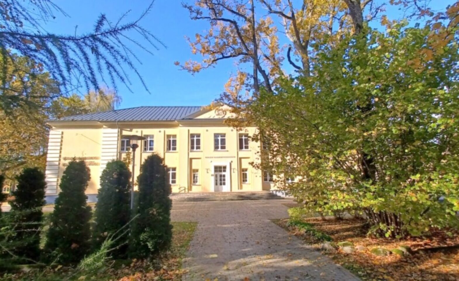 Gaiši dzeltena divstāvu ēka ar tumšu jumtu un baltiem logiem, ko ieskauj rudens koki ar dzeltenām un zaļām lapām, zem skaidrām zilām debesīm. Bruģēts celiņš ved uz galveno ieeju, un zemi klāj nokritušas lapas.