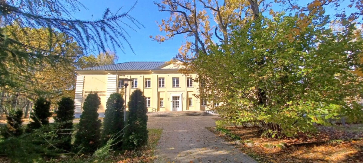 Gaiši dzeltena divstāvu ēka ar tumšu jumtu un baltiem logiem, ko ieskauj rudens koki ar dzeltenām un zaļām lapām, zem skaidrām zilām debesīm. Bruģēts celiņš ved uz galveno ieeju, un zemi klāj nokritušas lapas.