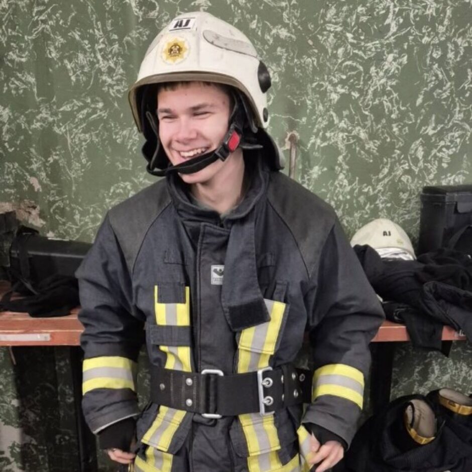 Smaidīgs jauns vīrietis ugunsdzēsēja uniformā ar baltu ķiveri, uz kuras rakstīts 