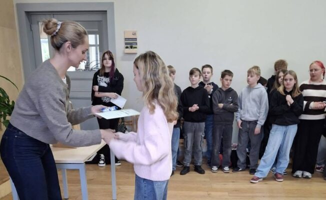 Sieviete pelēkā džemperī pasniedz sertifikātu vai balvu jaunai meitenei rozā džemperī. Fonā stāv citi skolēni, vērojot notiekošo gaišā telpā ar koka grīdu.