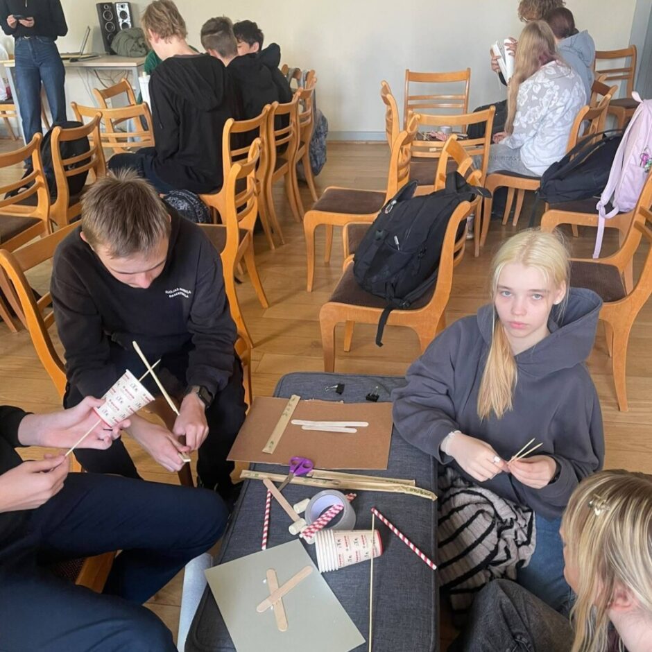 Grupa jauniešu strādā pie amatniecības projektiem telpā ar koka krēsliem; priekšplānā divi jaunieši būvē ar papīra glāzēm un koka kociņiem, kamēr citi sēž fonā.