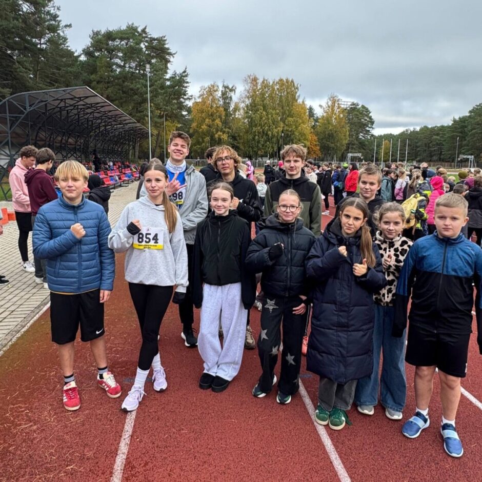 Bērnu un pusaudžu grupa, no kuriem daudzi saspiež dūres, stāv uz sarkanas skrejceliņa āra sporta pasākumā. Meitenei pelēkā džemperī ir numurs 