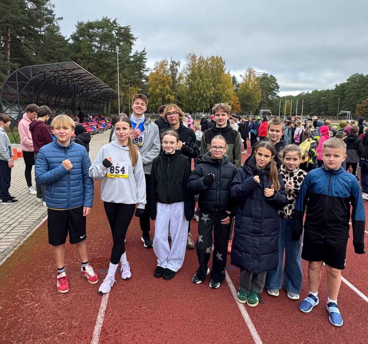 Bērnu un pusaudžu grupa, no kuriem daudzi saspiež dūres, stāv uz sarkanas skrejceliņa āra sporta pasākumā. Meitenei pelēkā džemperī ir numurs 