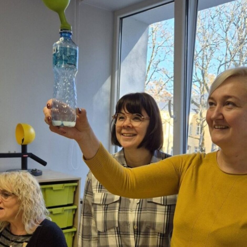 Trīs smaidīgas sievietes klasē. Viena sieviete dzeltenā džemperī tur augšup plastmasas pudeli, kurai uz kakla ir piepūsts zaļš balons, demonstrējot eksperimentu. Divas citas sievietes skatās un smaida. Uz galda priekšplānā redzami eksperimenta materiāli: etiķa pudele, mērtrauks ar baltu pulveri un citi piederumi.